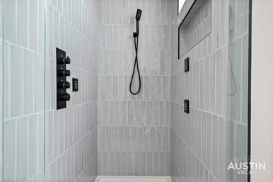 Sleek modern walk-in shower with glossy light-gray vertical subway tiles, matte black handheld shower head and control panel, recessed shelf niche and glass enclosure