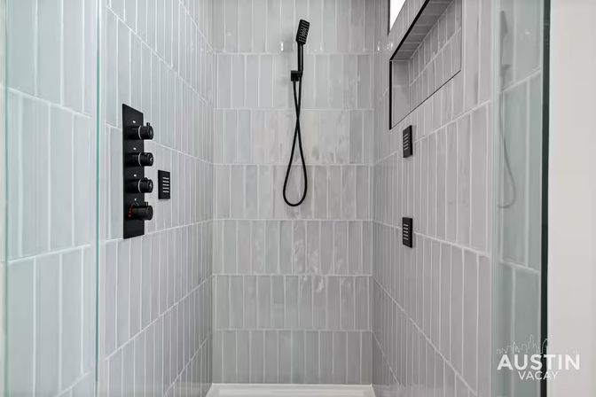 Sleek modern walk-in shower with glossy light-gray vertical subway tiles, matte black handheld shower head and control panel, recessed shelf niche and glass enclosure