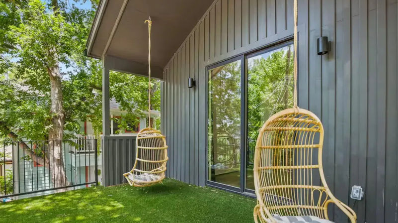 Covered modern residential balcony with artificial turf, two hanging rattan egg chairs, dark vertical siding and sliding glass door overlooking leafy trees