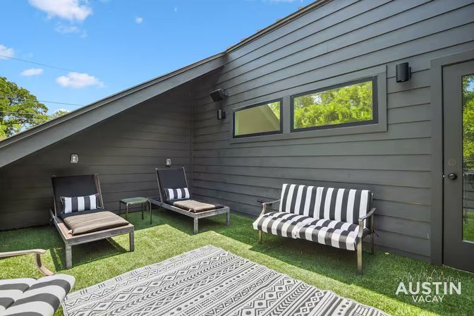Sunny rooftop patio with modern gray siding, striped outdoor sofa and chaise loungers on artificial turf and a geometric rug