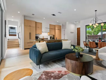 Bright modern open-plan living room and kitchen with navy sofa, round wooden coffee table, light oak cabinets, dining area and sliding glass doors to a leafy backyard.