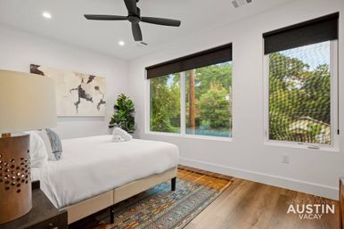 Bright modern bedroom in Austin with large windows overlooking trees, white bed with rolled towel, black ceiling fan, abstract wall art, potted plant, wood floors and colorful patterned rug.