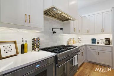 Bright modern white kitchen with a stainless steel professional gas range, swing-out pot filler, white subway-tile backsplash, brass cabinet pulls, spice carousel and olive oil bottles, coffee station, and warm hardwood floors.