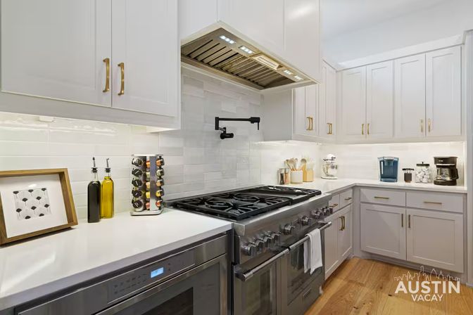 Bright modern white kitchen with a stainless steel professional gas range, swing-out pot filler, white subway-tile backsplash, brass cabinet pulls, spice carousel and olive oil bottles, coffee station, and warm hardwood floors.