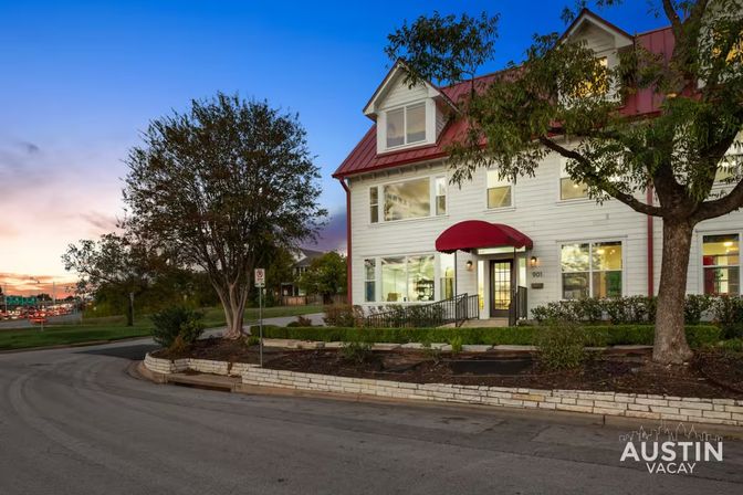 Two-story white building with a red metal roof and red awning lit at dusk, framed by trees along a curved street and a colorful sunset sky in Austin.