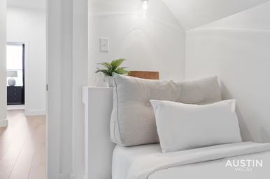 Cozy minimalist bedroom nook with plush white pillows and duvet, a small potted fern on a ledge, soft overhead light, and a hardwood-floored hallway visible through an open doorway.