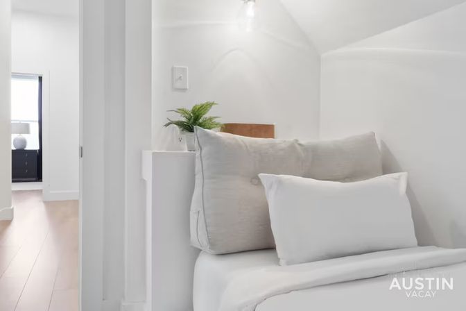 Cozy minimalist bedroom nook with plush white pillows and duvet, a small potted fern on a ledge, soft overhead light, and a hardwood-floored hallway visible through an open doorway.