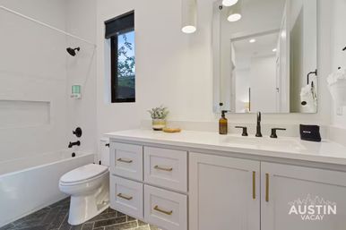 Bright modern bathroom with white tub/shower, toilet, white vanity with gold drawer pulls, matte-black faucet and shower fixtures, gray herringbone tile floor, small window, potted plant and large mirror.