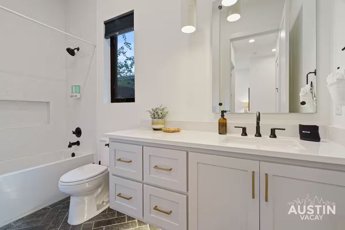 Bright modern bathroom with white tub/shower, toilet, white vanity with gold drawer pulls, matte-black faucet and shower fixtures, gray herringbone tile floor, small window, potted plant and large mirror.