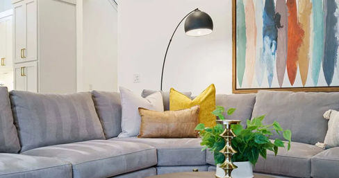 Cozy modern living room with a light-gray sectional sofa, mustard and tan throw pillows, arched black floor lamp, colorful abstract wall art, and a coffee table featuring a potted green plant and brass candleholder.