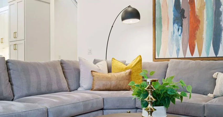 Cozy modern living room with a light-gray sectional sofa, mustard and tan throw pillows, arched black floor lamp, colorful abstract wall art, and a coffee table featuring a potted green plant and brass candleholder.