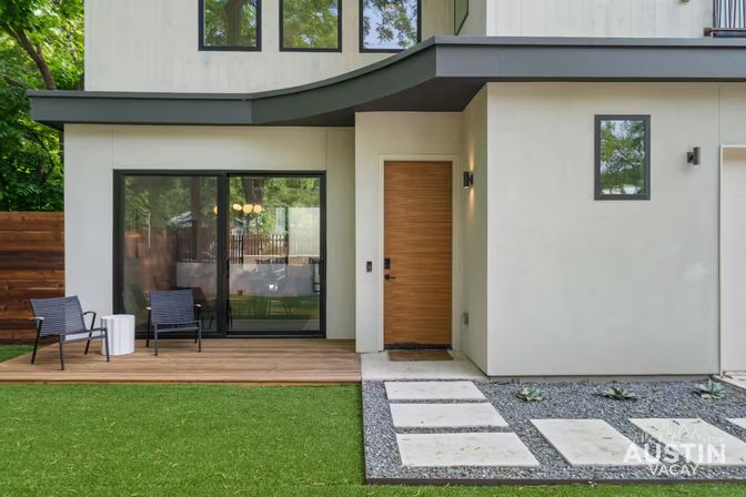 Modern minimalist home exterior with wooden front door, large sliding glass patio door, small wood deck and chairs, artificial turf lawn and concrete stepping stones.