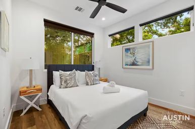 Modern Austin vacation rental bedroom with a queen bed in crisp white linens, decorative pillows and a rolled towel, dark upholstered headboard, wooden nightstands, black ceiling fan, high windows with leafy tree views and hardwood floors.