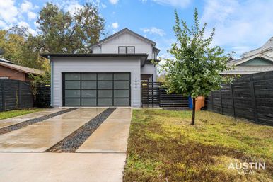 Modern two-story gray home with frosted glass-panel garage door, wet concrete driveway with gravel strips, small front lawn and lone tree, black privacy fence and cloudy sky.