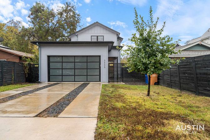 Modern two-story gray home with frosted glass-panel garage door, wet concrete driveway with gravel strips, small front lawn and lone tree, black privacy fence and cloudy sky.