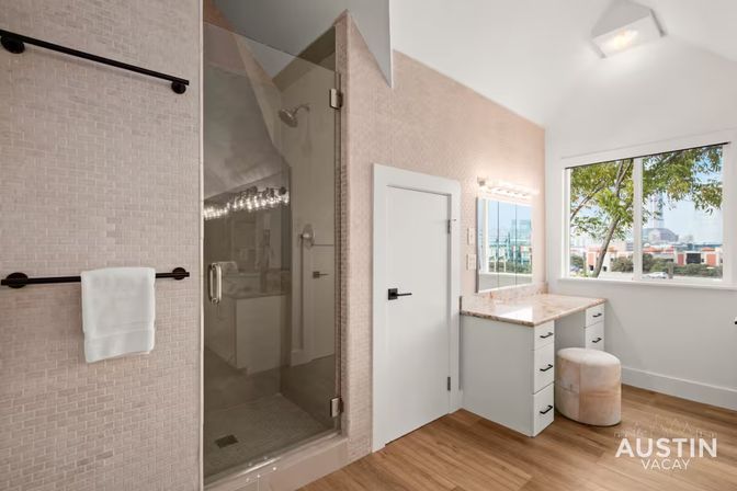 Sunlit modern bathroom with frameless glass shower, towel bar with white towel, marble-topped vanity and cushioned stool, hardwood floor and large window framing trees and a distant urban skyline.