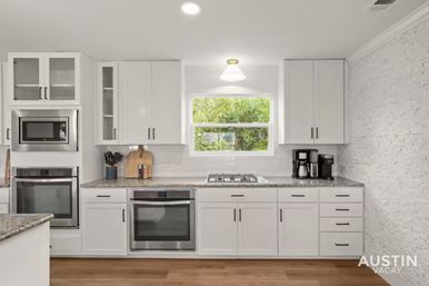 Bright modern white kitchen in a vacation-rental style: stainless steel ovens and microwave, gas cooktop beneath a window with greenery view, granite countertops, coffee maker, wood cutting boards, and textured white brick accent wall.
