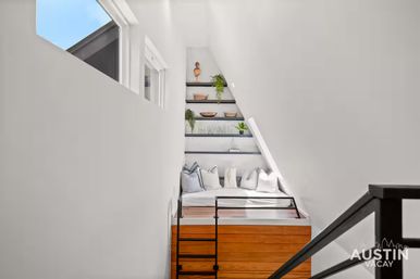 Cozy modern loft sleeping nook with white angled walls, built-in wooden platform bed and black ladder, plush pillows, floating shelves with plants and decor, and a skylight window letting in blue sky light.