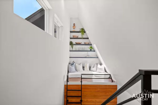 Cozy modern loft sleeping nook with white angled walls, built-in wooden platform bed and black ladder, plush pillows, floating shelves with plants and decor, and a skylight window letting in blue sky light.