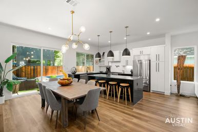 Bright modern Austin, TX open-plan kitchen and dining area with a wooden table and gray chairs, black island with wooden bar stools, brass and pendant lighting, and sliding glass doors overlooking a fenced backyard pool.
