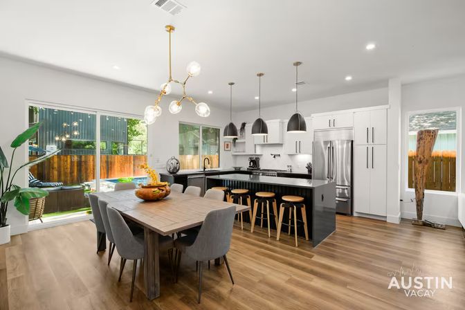 Bright modern Austin, TX open-plan kitchen and dining area with a wooden table and gray chairs, black island with wooden bar stools, brass and pendant lighting, and sliding glass doors overlooking a fenced backyard pool.