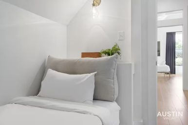 Cozy minimalist bedroom nook in a bright vacation rental with white linens and gray pillows, exposed bulb sconce, potted fern on a shelf, and a view into a sunlit room with hardwood floors.