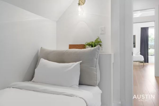 Cozy minimalist bedroom nook in a bright vacation rental with white linens and gray pillows, exposed bulb sconce, potted fern on a shelf, and a view into a sunlit room with hardwood floors.