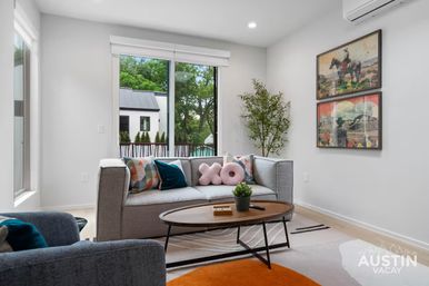 Sunlit modern Austin living room in a vacation rental — gray sofa with geometric and plush pink pillows, round wooden coffee table, potted plant, framed western art, and large window with treetop view.