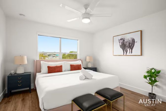Bright modern bedroom with king bed dressed in white linens and coral pillows, navy nightstands with lamps, ceiling fan, buffalo wall art, two black ottomans and a potted plant