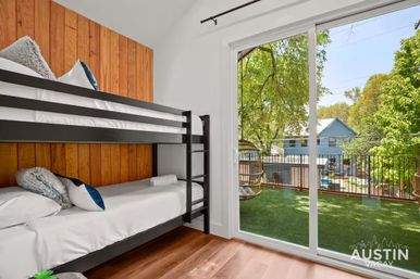 Cozy modern bunk bedroom with black metal frame, white linens and wood-paneled accent wall opening to a sliding-glass door and balcony with hanging chair and leafy neighborhood view.