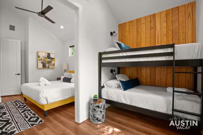 Bright Austin vacation rental bedroom with a yellow platform queen bed, black metal bunk beds against wood-paneled accent wall, hardwood floors, ceiling fan, patterned rug and a small stump side table with a plant.