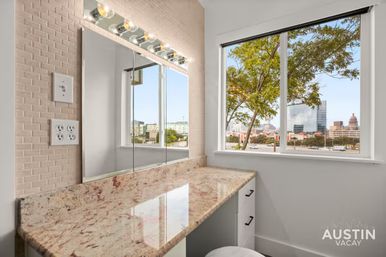 Sunlit dressing vanity with speckled marble countertop, Hollywood-style lightbulb mirror and drawers, window framing a leafy tree and downtown Austin skyline.