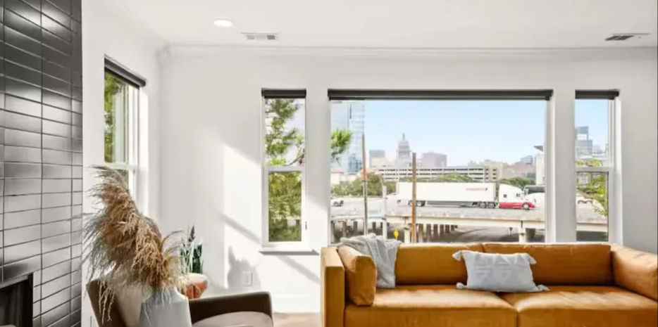 Bright modern living room with caramel leather sofa, large windows framing an urban highway and skyline, tiled fireplace and indoor plants.