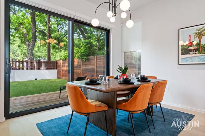 Bright modern dining room with wooden table set for four, orange velvet chairs, globe pendant light, and sliding glass doors opening to a fenced backyard with green lawn and trees