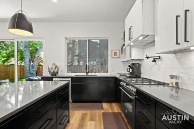 Bright modern Austin kitchen with black lower cabinets and white uppers, large quartz island, stainless gas range and coffee maker, pendant light, window and sliding glass door looking onto a tree-lined backyard with wooden fence.