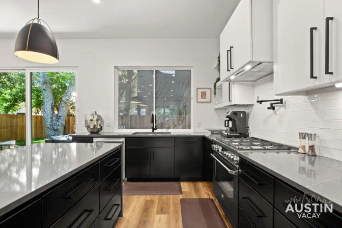 Bright modern Austin kitchen with black lower cabinets and white uppers, large quartz island, stainless gas range and coffee maker, pendant light, window and sliding glass door looking onto a tree-lined backyard with wooden fence.
