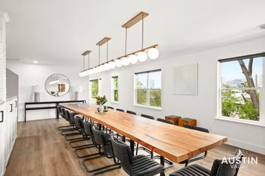 Bright modern conference room with a long live-edge wood table, black leather chairs, linear globe pendant lights, hardwood floors, white walls, windows showing a tree-lined city view, and a round mirror with a console table at the far end.