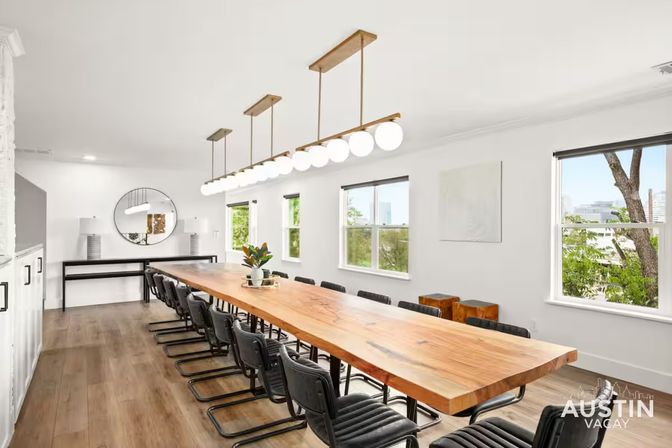 Bright modern conference room with a long live-edge wood table, black leather chairs, linear globe pendant lights, hardwood floors, white walls, windows showing a tree-lined city view, and a round mirror with a console table at the far end.