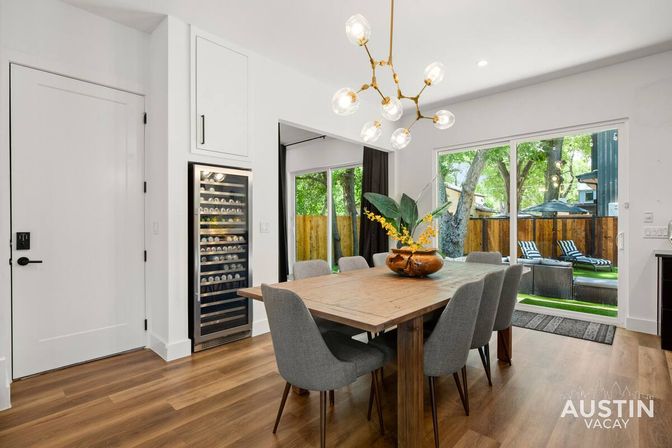 Bright modern Austin dining room — wooden farmhouse table with six gray chairs, globe chandelier, built-in wine fridge, sliding glass doors to a tree-shaded backyard with lounge chairs