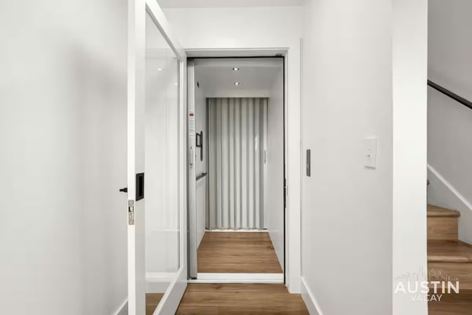 Modern residential elevator with open glass door and folding accordion gate, white walls and light wood floors in a bright hallway beside wooden stairs