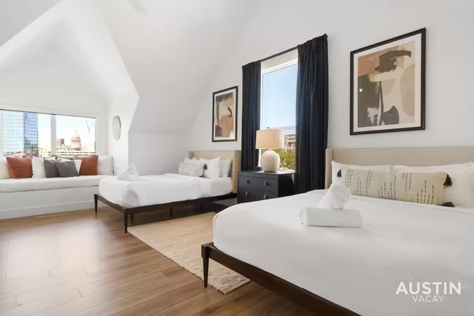 Bright modern guestroom with vaulted ceiling, two white beds with decorative pillows, a built-in window seat overlooking a downtown skyline, hardwood floors, black curtains, bedside lamp and abstract wall art.