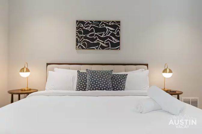 Modern urban hotel-style king bed with crisp white linens and rolled towel, three gray patterned accent pillows, brass globe bedside lamps on wooden tables, and abstract black-and-white artwork above the headboard.