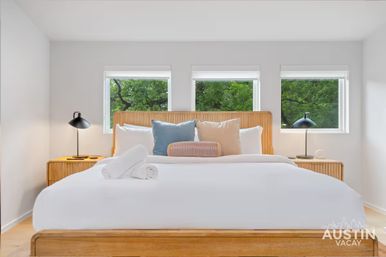 Bright modern vacation-style bedroom with a king bed, crisp white linens, wooden headboard, colorful throw pillows, bedside lamps and three windows framing green trees.