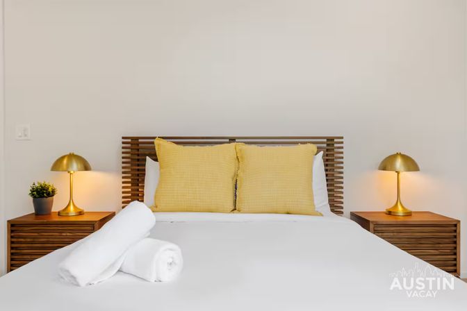 Minimalist bedroom with a wooden slatted headboard, two cheerful yellow pillows, gold bedside lamps on matching wooden nightstands, and rolled white towels on crisp white bedding.
