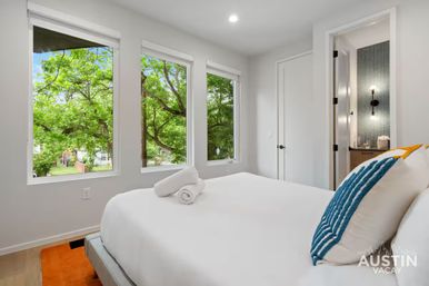 Sunlit modern bedroom with crisp white bed, rolled towel and blue-striped pillow, three large windows framing leafy green trees and a visible en-suite bathroom.