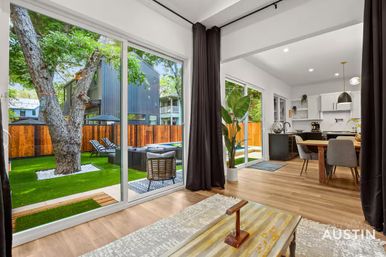 Bright modern open-concept living room and kitchen with wood floors and dark curtains, wide sliding glass doors opening to a private fenced backyard patio with a large mature tree, lounge seating and artificial turf.