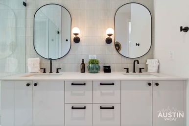 Sleek modern double-sink bathroom vanity with white cabinets, matte black faucets and hardware, two arched mirrors, round wall sconces, vertical textured tile backsplash, and a small green plant on the countertop.