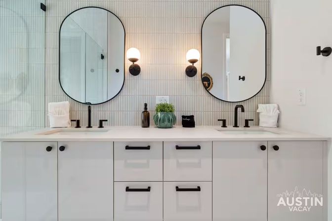 Sleek modern double-sink bathroom vanity with white cabinets, matte black faucets and hardware, two arched mirrors, round wall sconces, vertical textured tile backsplash, and a small green plant on the countertop.