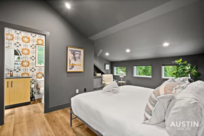 Cozy modern loft bedroom with dark gray sloped ceiling, recessed lights, white linens and patterned throw pillows, rolled towel on the bed, low windows with green views, potted plant and wood floors; open doorway shows a bathroom with orange-gray patterned tile, round mirror and light wood vanity.