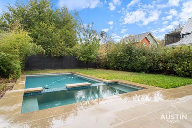 Residential backyard with a rectangular lap pool and attached spa, wet stone decking, green lawn and tall privacy hedges, nearby rooftops and a blue sky with scattered clouds.